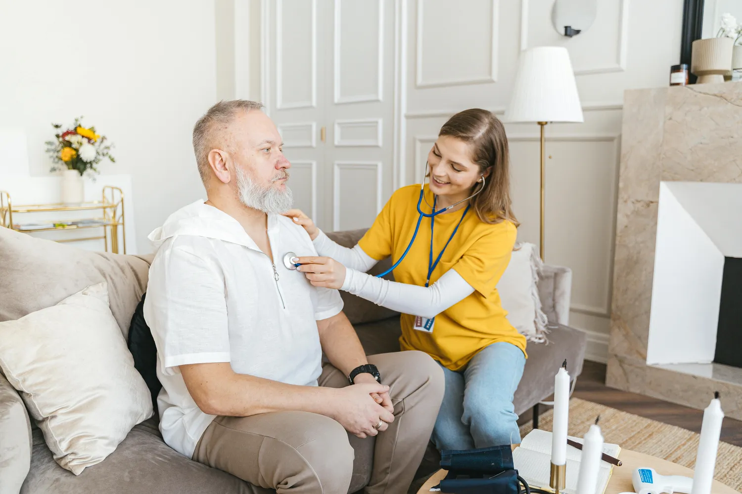 FamilyCare Leipzig UG – Pflegeleistungen für alle Altersgruppen – Eine Pflegekraft benutzt ein Stethoskop bei einer Hausuntersuchung eines älteren Menschen in einem gemütlichen Wohnzimmer.